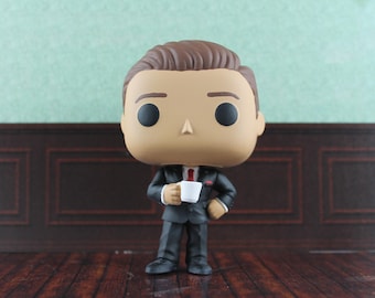 May include: A vinyl collectible figure featuring a character with brown hair, wearing a dark grey suit with a red tie and a white pocket square. The figure holds a small white coffee mug in one hand and stands in a formal pose with a large head.