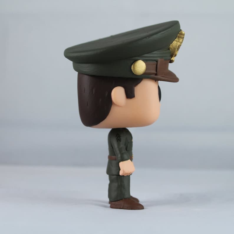 Peut inclure: Figurine d'officier militaire vue de c&ocirc;t&eacute;. La figurine porte un uniforme vert fonc&eacute;, une ceinture marron et des chaussures marron. Le chapeau de l'officier est vert fonc&eacute; avec un bord marron et des d&eacute;tails dor&eacute;s.