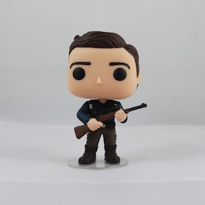 May include: A vinyl figure of a person with brown hair, black eyes, and a neutral expression. The figure is wearing a dark blue shirt, brown pants, and black boots. It is holding a brown rifle. The figure is standing on a clear plastic base.