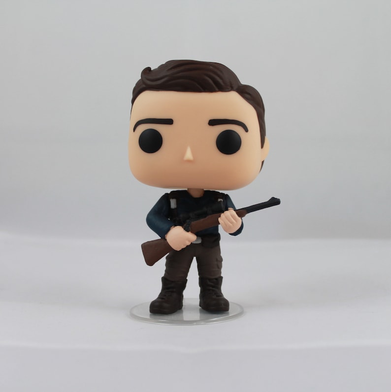 May include: A vinyl figure of a person with brown hair, black eyes, and a neutral expression. The figure is wearing a dark blue shirt, brown pants, and black boots. It is holding a brown rifle. The figure is standing on a clear plastic base.
