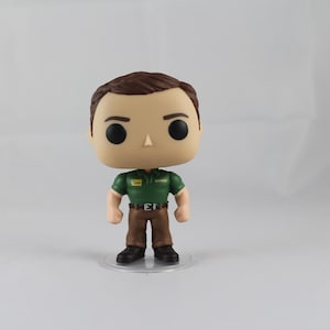 Puede incluir: Una figura de vinilo pop verde y marrón de un hombre que lleva una camisa verde con una etiqueta de nombre que dice "Greg" y pantalones marrones. La figura está de pie sobre una base de plástico transparente.