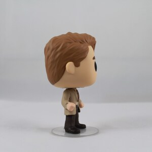 May include: A Funko Pop vinyl figure of a man with brown hair, wearing a tan jacket, dark trousers, and black shoes. The figure stands on a clear, circular base. The figure has a large head and black eyes.