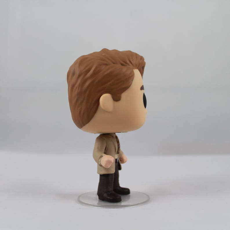 May include: A Funko Pop vinyl figure of a man with brown hair, wearing a tan jacket, dark trousers, and black shoes. The figure stands on a clear, circular base. The figure has a large head and black eyes.