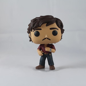 May include: A vinyl figure of a man with dark brown hair and a moustache, wearing a maroon shirt, dark blue trousers, and brown shoes. The figure has a watch on its left wrist and stands on a clear base.