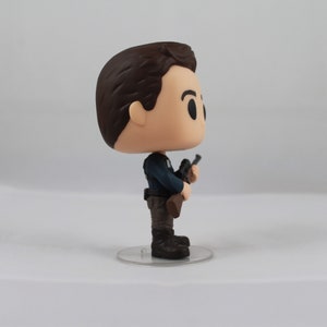 May include: A vinyl figure of a man in a dark blue uniform, holding a rifle. The figure has a large head and small body, with brown hair and black boots. It stands on a clear plastic base.