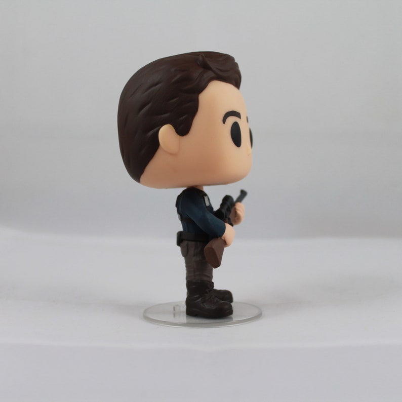 May include: A vinyl figure of a man in a dark blue uniform, holding a rifle. The figure has a large head and small body, with brown hair and black boots. It stands on a clear plastic base.