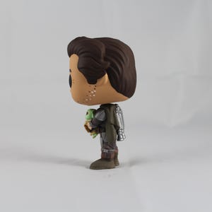 May include: A vinyl figure of a character from a science fiction series. The figure depicts a man in a silver and green suit holding a small green creature. The figure has brown hair and a tan face. The figure is standing on a white surface.