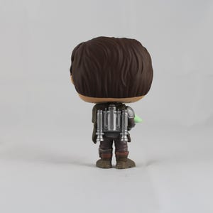 May include: A vinyl figure of a character from the Star Wars franchise, viewed from the back. The figure has brown hair, a silver jetpack, and brown and gray clothing. A small green object is visible on the right side of the figure.