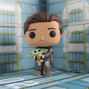 May include: A Funko Pop vinyl figure of the Mandalorian holding Grogu, also known as Baby Yoda. The figure has a tan complexion, brown hair, and a mustache. Grogu is green with large ears and is wearing a brown and black outfit. The background is a blue and white interior.