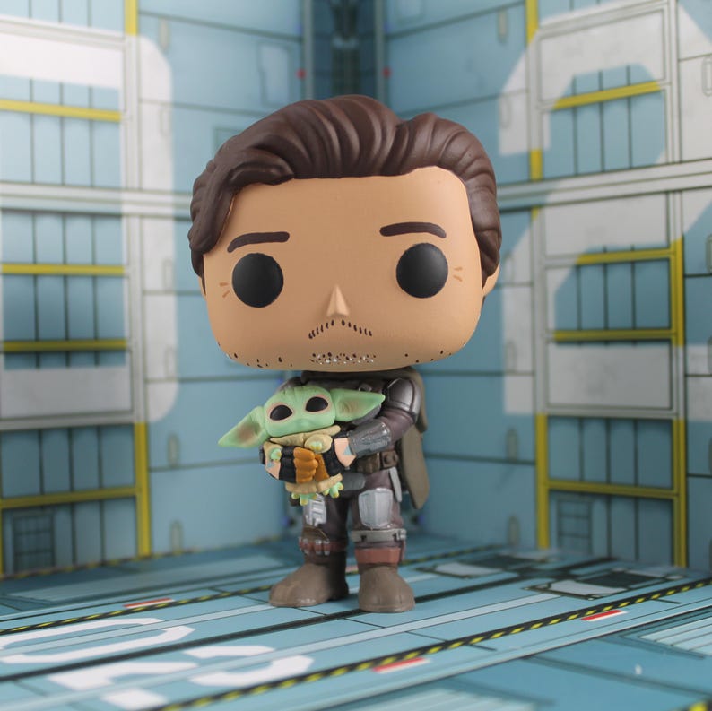 May include: A Funko Pop vinyl figure of the Mandalorian holding Grogu, also known as Baby Yoda. The figure has a tan complexion, brown hair, and a mustache. Grogu is green with large ears and is wearing a brown and black outfit. The background is a blue and white interior.