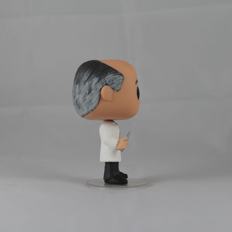 Puede incluir: Una figura de vinilo blanca y negra de un hombre con cabello gris que lleva una bata de laboratorio blanca y sostiene un peque&ntilde;o objeto plateado. La figura est&aacute; de pie sobre una base de pl&aacute;stico transparente.