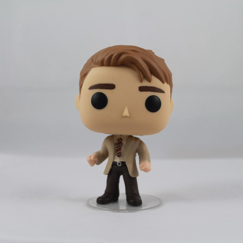 May include: A Funko Pop vinyl figure of a man with brown hair, wearing a tan jacket, brown trousers, and a patterned tie. The figure has large black eyes and is standing on a clear plastic base. The figure is approximately 10 cm tall.