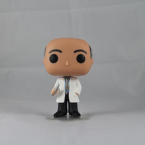 Puede incluir: Una figura de vinilo blanca y marr&oacute;n de un hombre calvo que lleva una bata de laboratorio blanca, una corbata a rayas azul y marr&oacute;n y pantalones negros. Sostiene un peque&ntilde;o objeto blanco en su mano derecha. La figura est&aacute; de pie sobre una base de pl&aacute;stico transparente.