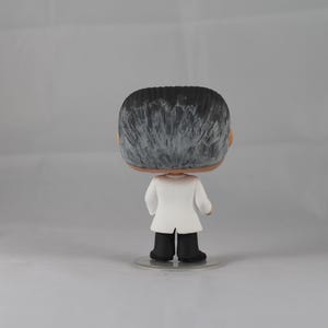 Puede incluir: Una figura de vinilo en blanco y negro de una persona que lleva una bata de laboratorio blanca. La figura tiene una base negra.