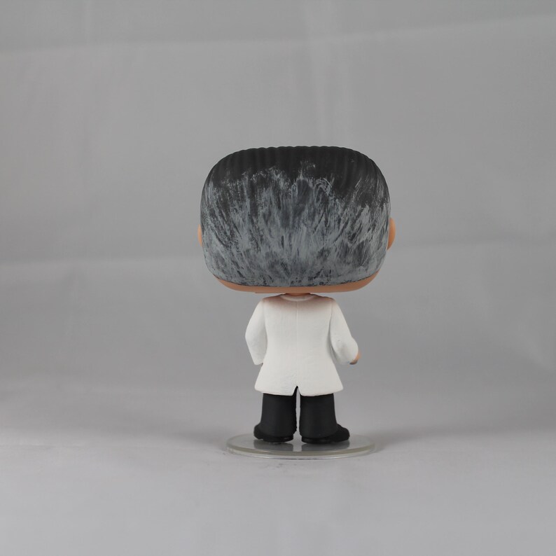 Puede incluir: Una figura de vinilo en blanco y negro de una persona que lleva una bata de laboratorio blanca. La figura tiene una base negra.