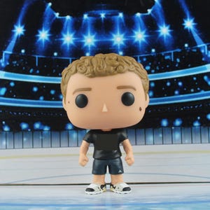Op de afbeelding: Een vinyl verzamelfiguur van een persoon met lichtbruin krullend haar, gekleed in een zwart t-shirt, een donkere korte broek met een klein wit logo op de pijp en witte sneakers met zwarte accenten. Het figuurtje heeft een groot hoofd en zwarte ogen.
