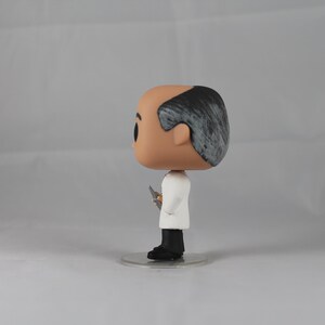 Puede incluir: Una figura de vinilo blanca y gris de un hombre con cabello gris, que lleva una bata de laboratorio blanca y sostiene un peque&ntilde;o objeto plateado en su mano derecha. La figura est&aacute; de pie sobre una base de pl&aacute;stico transparente.