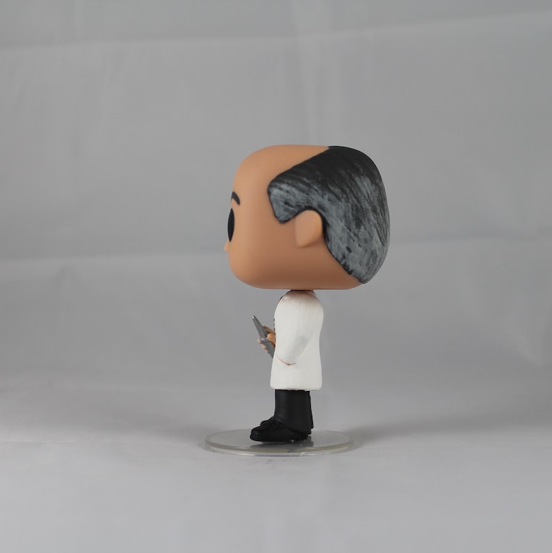Puede incluir: Una figura de vinilo blanca y gris de un hombre con cabello gris, que lleva una bata de laboratorio blanca y sostiene un peque&ntilde;o objeto plateado en su mano derecha. La figura est&aacute; de pie sobre una base de pl&aacute;stico transparente.