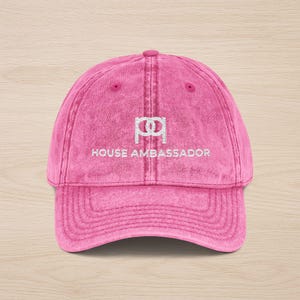 May include: A pink baseball cap with a curved brim. The cap features the embroidered text "HOUSE AMBASSADOR" in white, along with a stylized logo above the text. The cap has a vintage, washed look.