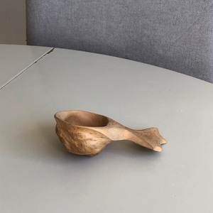 Può includere: Una tazza kuksa in legno fatta a mano con finitura naturale. La tazza presenta una forma rotonda e un manico intagliato. La venatura del legno è visibile, mostrando la bellezza naturale. Questa tazza rustica è perfetta per il campeggio.