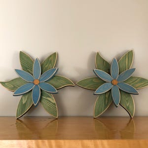 May include: Two decorative wooden flower wall hangings, each featuring a layered design with six large green-stained leaves as a base and six smaller blue-stained petals on top, centred with a small circular natural wood grain accent piece.