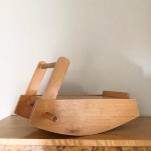 May include: A light-colored wooden rocking cradle with a smooth finish. The structure features curved side panels, a horizontal handle bar at the top, and a small cylindrical peg protruding from the side. The design is simple and functional.