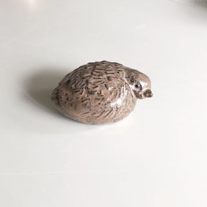 May include: A small, rounded ceramic bird figurine with a glossy, light brown finish. The surface features dark, etched, dash-like markings that mimic feathers across the body, with a smooth head, a small beak, and a single visible dark eye.