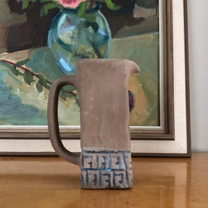 May include: A tall, cylindrical ceramic pitcher with a matte tan finish on the upper portion and a textured, grid-like pattern in blue and tan on the base. The pitcher features a curved handle on the left side and a small pouring spout at the top.