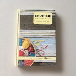 May include: A hardcover book titled "TRASMATTOR OCH ANDRA MATTOR" with a striped cover in shades of brown, gray, and white. The book features a basket filled with colorful yarn and ribbons. The text "EN VÄVBOK FRÅN ICA" is also visible.