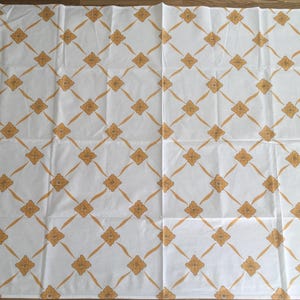 May include: White fabric with a repeating pattern of gold-colored floral and diamond shapes. The design is evenly spaced across the fabric, creating a symmetrical and visually appealing pattern. The fabric appears to be a tablecloth or similar textile.