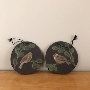 Könnte beinhalten: Zwei kreisförmige Wandplaketten aus dunkelbrauner Keramik, die jeweils einen handbemalten braunen Vogel auf einem grünen Blattzweig zeigen. Jede Plakette ist mit einer schwarzen Schnurschlaufe an der Oberseite zum Aufhängen versehen.