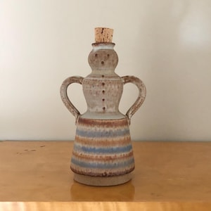 May include: A ceramic bottle with a cork stopper, featuring a rounded neck and two curved handles. The upper portion is speckled beige, while the lower body displays horizontal stripes in alternating shades of blue, brown, and cream glaze.