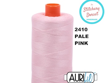 May include: A spool of pale pink Aurifil thread, color 2410, with an orange top and base. The thread is tightly wound, and the label includes "The Stitchery Dorset Handmade".