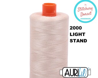 May include: A spool of 2000 Light Stand Aurifil thread in a light beige color. The spool has a red plastic top and bottom. The text "The Stitchery Dorset Handmade" is in a circle on the right side of the image. The text "2000 Light Stand" is below the circle. The text "Aurifil" is in a blue rectangle on the bottom right side of the image.