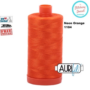 May include: A vibrant neon orange thread spool, marked "Neon Orange 1104." The thread is neatly wrapped around a red plastic spool. The brand name "AuriFil" is displayed. The Stitchery Dorset handmade logo is in the upper right corner.