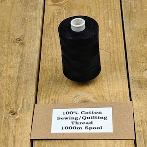 Puede incluir: Una bobina negra de hilo de coser y acolchar 100% algodón. La bobina tiene un núcleo blanco y está etiquetada como 1000m. La bobina está sobre una superficie de madera.