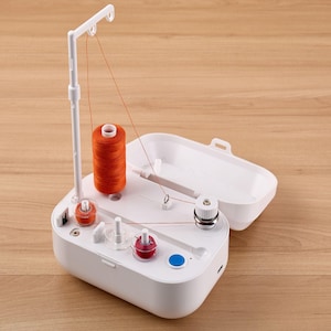 May include: A white sewing machine with an open lid, featuring a spool of orange thread and several smaller spools in red and white. A blue button is visible on the machine's surface. The machine is set on a wooden surface.