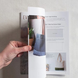 May include: Open book with instructions on how to make a tote bag. The book shows a close-up of a blue denim tote bag with a brown leather handle and a smaller pocket on the front. The text on the page reads "Prepare: Apply interfacing" and "Step 1: Make Front Exterior Pocket".