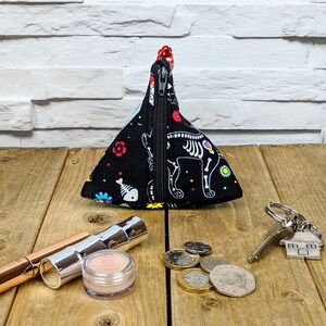 May include: A black triangular zippered pouch with a colorful skeleton and floral print. The pouch is sitting on a wooden surface with a silver key chain and coins.