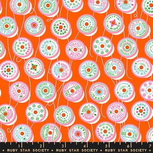 Op de afbeelding: Oranje stof met een herhalend patroon van roze, groene en witte garenklosjes. Elke klos heeft tekst zoals "Tightly Wound" en "Sew With Love". De stof is van Ruby Star Society.