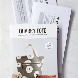 May include: A sewing pattern titled "QUARRY TOTE" with a flat bottom tote design in two sizes, plus a bonus zippered pouch. The pattern cover shows a tote bag with white circular patterns on a gray background, and a small orange pouch.