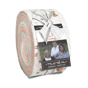 Könnte beinhalten: Gerolltes Stoffbündel in Pastelltönen. Das Gewebe ist mit einem weißen Band und einem Etikett mit dem Logo "Moda Fabrics + Supplies" und einem Foto von drei Frauen umwickelt. Der Stoff hat ein sich wiederholendes Muster aus kleinen Bildern.