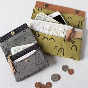 May include: Two fabric wallets, one in a black and white geometric pattern and the other in a green and white pattern with black arches. Both wallets are open and show cash and cards inside. There are also several coins scattered on the white surface.