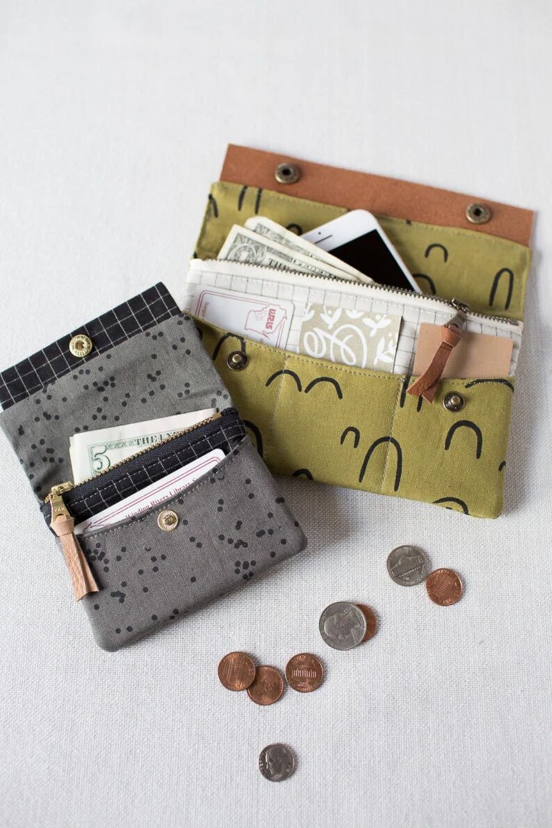 May include: Two fabric wallets, one in a black and white geometric pattern and the other in a green and white pattern with black arches. Both wallets are open and show cash and cards inside. There are also several coins scattered on the white surface.