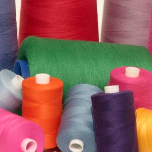 Könnte beinhalten: Eine Sammlung farbenfroher Garnrollen in verschiedenen Farbtönen, darunter Rot, Grün, Pink, Blau, Orange, Lila und Gelb. Die Spulen sind eng gewickelt und haben weiße Kerne, ideal für Näh- und Bastelprojekte.