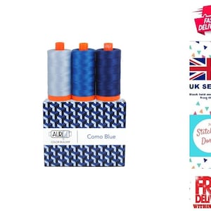 May include: Three spools of thread in varying shades of blue, ranging from light to dark, are positioned on a rectangular box. The box features a geometric pattern in blue and white, with the text AuriFil Color Builder Como Blue printed on it.