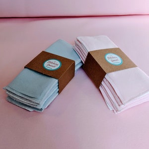 Puede incluir: Dos pilas de almohadillas de desmaquillaje reutilizables, una azul y otra rosa, con una etiqueta de papel marr&oacute;n que dice "Stitchery Dorset".