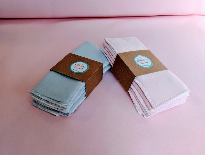 Puede incluir: Dos pilas de almohadillas de desmaquillaje reutilizables, una azul y otra rosa, con una etiqueta de papel marr&oacute;n que dice "Stitchery Dorset".
