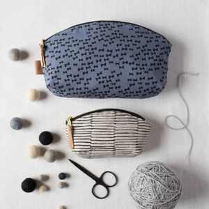 May include: Two fabric zippered pouches, one with a black bow pattern on a blue background and the other with black stripes on a white background. A ball of gray yarn, a pair of black scissors, and a scattering of gray and beige felt balls are on a white surface.