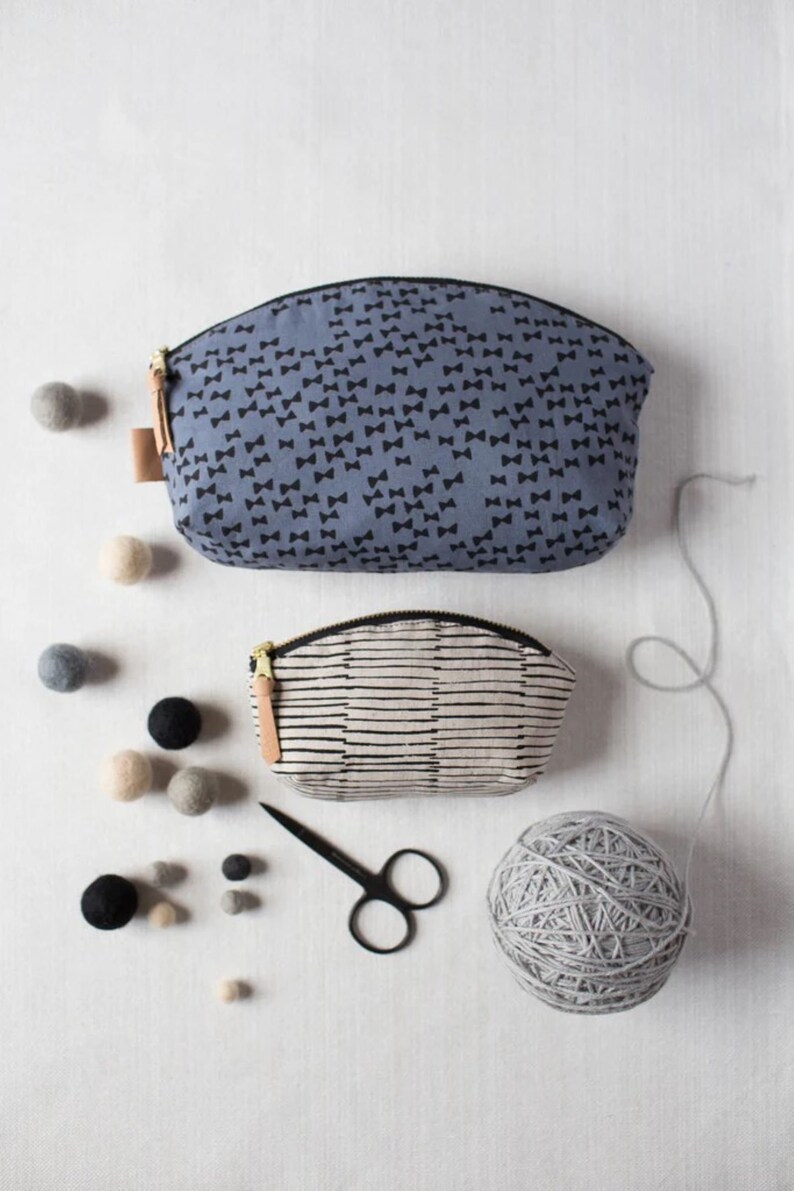 May include: Two fabric zippered pouches, one with a black bow pattern on a blue background and the other with black stripes on a white background. A ball of gray yarn, a pair of black scissors, and a scattering of gray and beige felt balls are on a white surface.